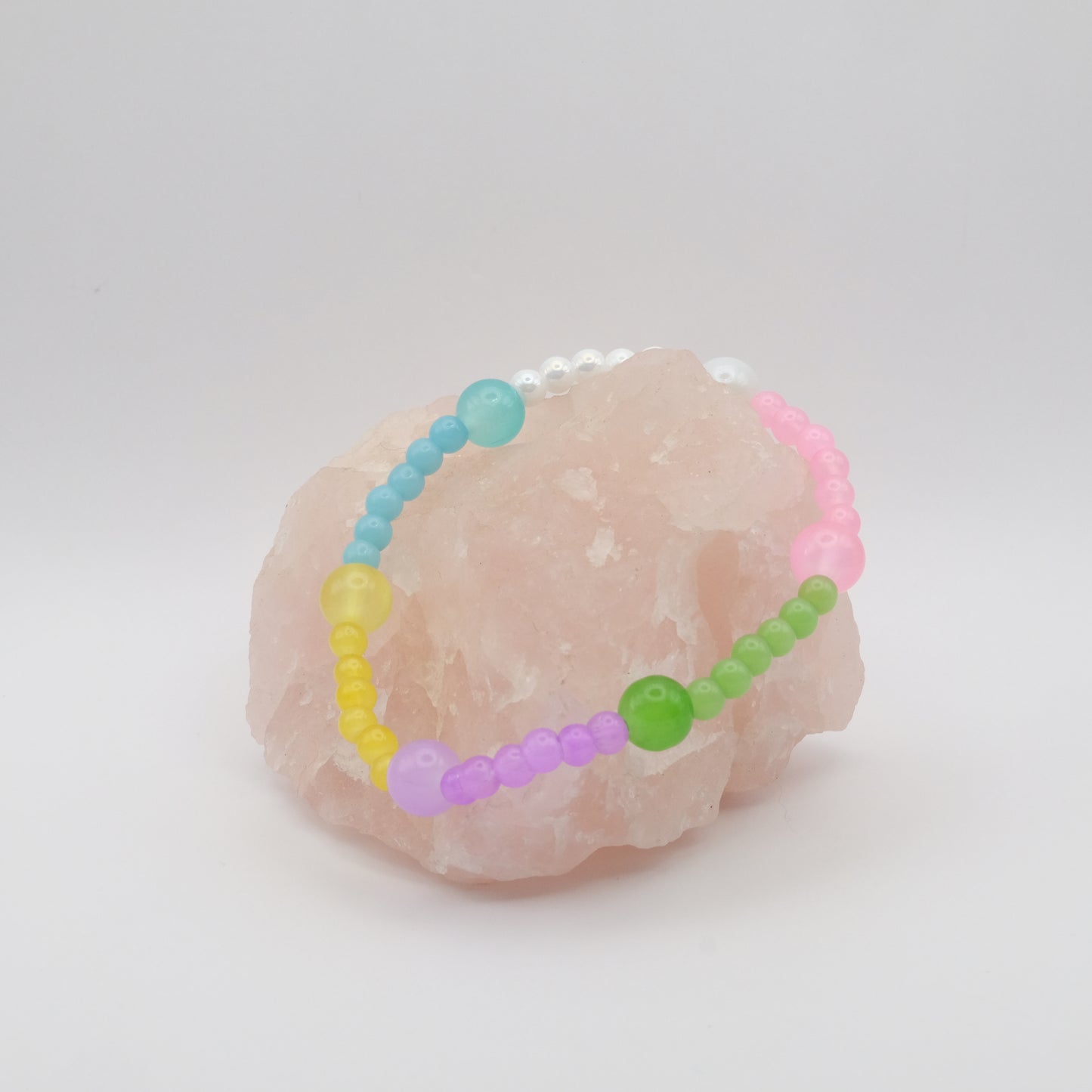 A single pastel coloured elastic beaded bracelet in pink, green, blue, purple, yellow and white sitting on a rose quartz rock with a white background