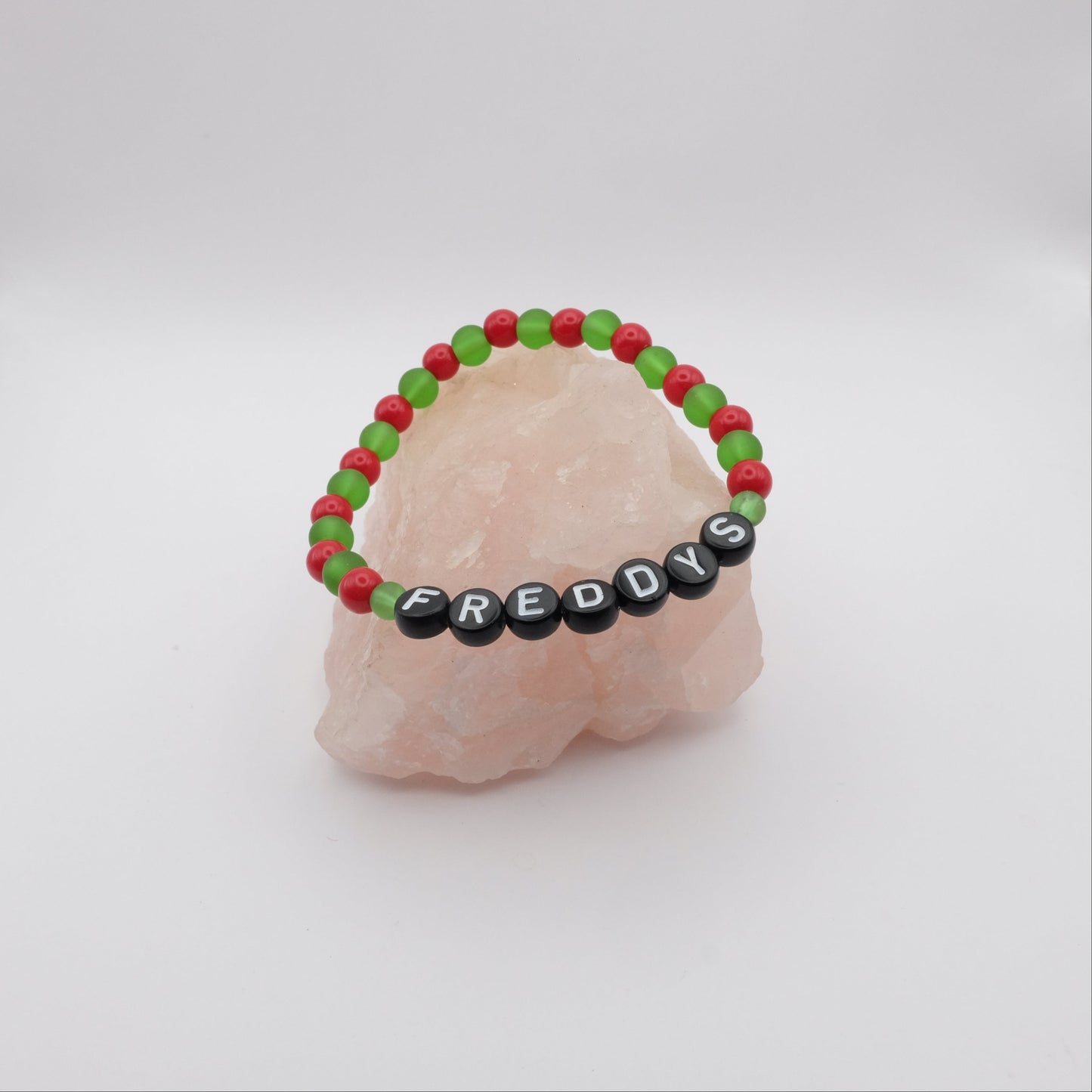 Bracelet with red and green beads and black beads spelling 'FREDDYS' on a light pink stone background