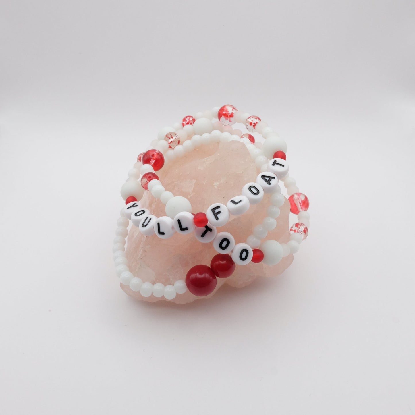 Beaded bracelets with red and white beads on a pink stone with 'YOU'LL FLOAT TOO' text.