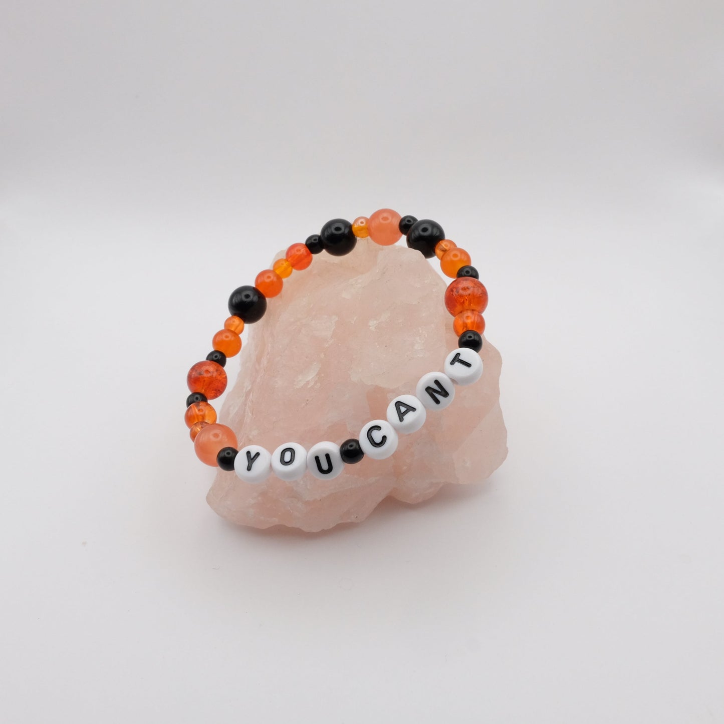 Black and orange bracelet with YOU CANT spelled out in white letter beads, placed on a pink stone