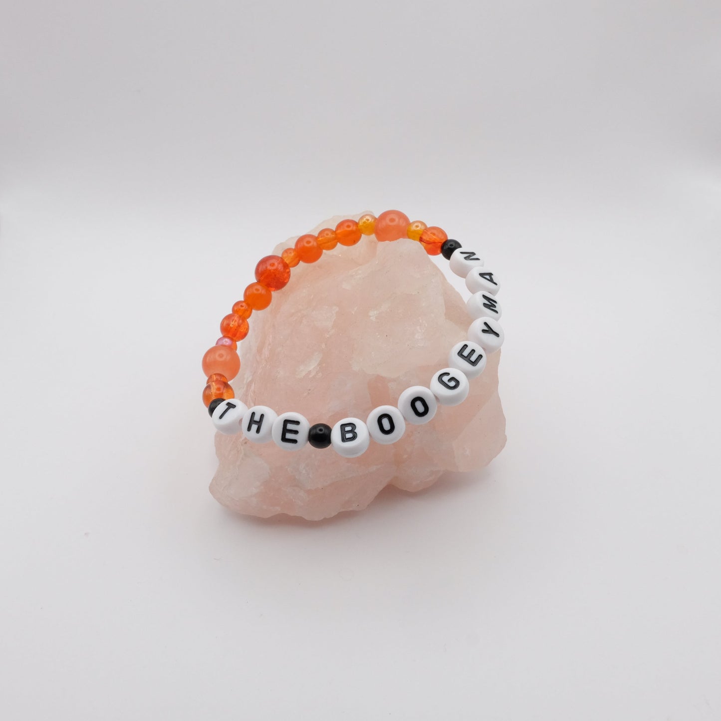 Black and orange bracelet with letter beads spelling THE BOOGEYMAN sitting on a rose quartz rock against a white background
