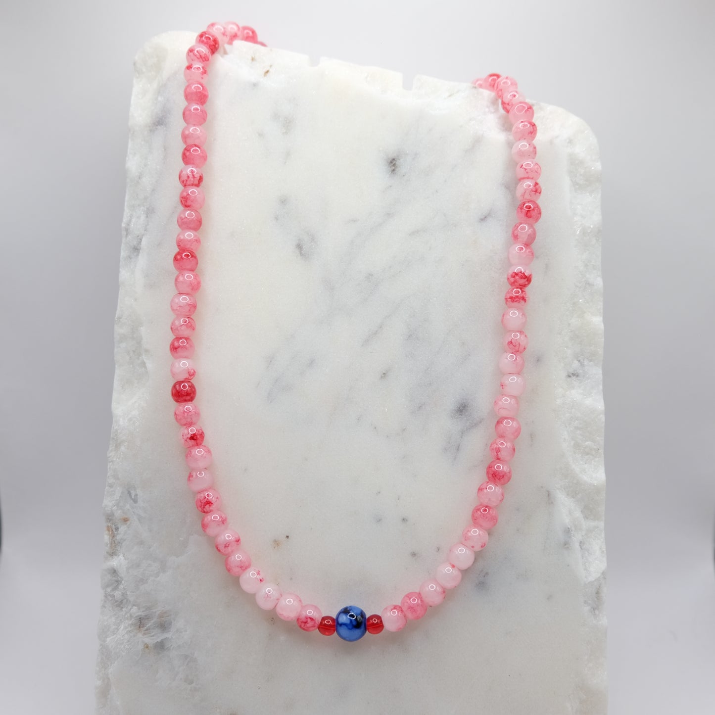 A beaded necklace composed of 6mm red and white marbled glass beads - featuring an 8mm blue and white marbled centre bead with small red accents, shown on a white marble background.