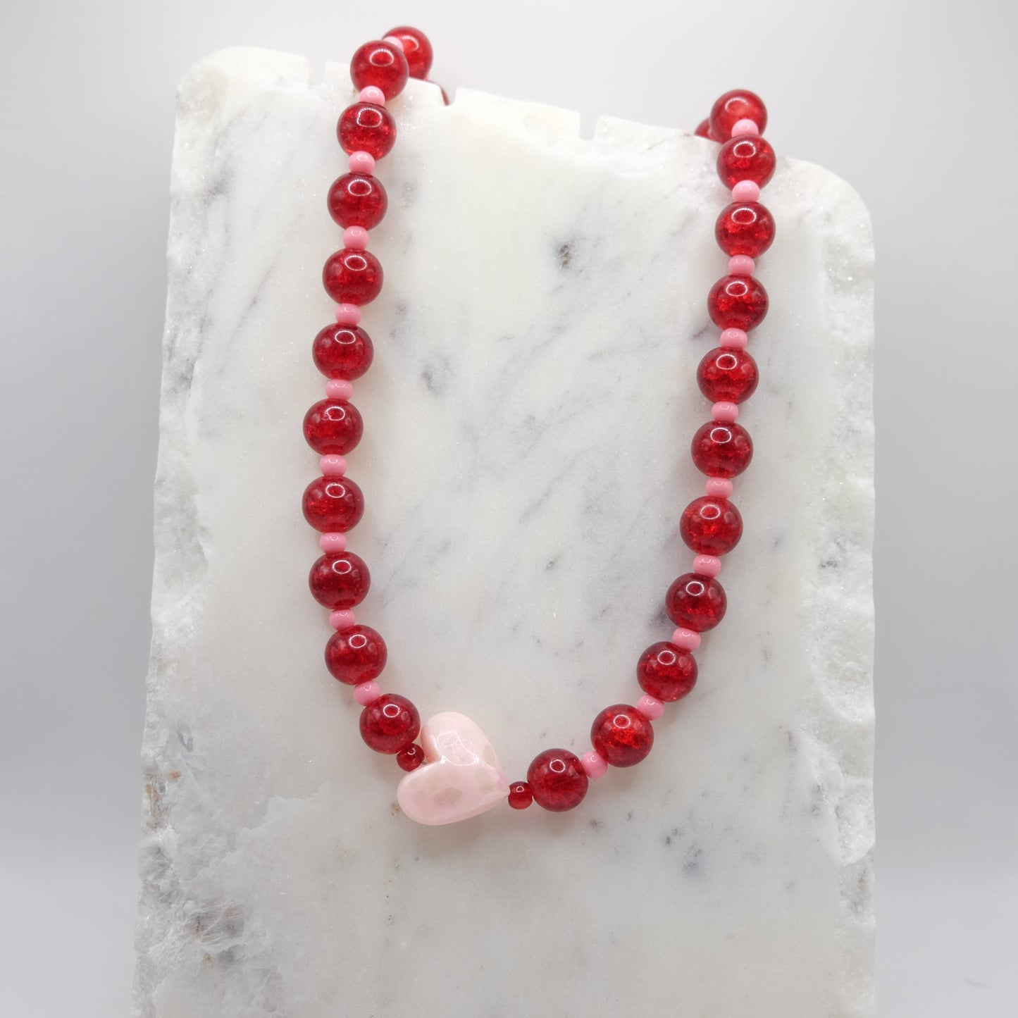 A beaded necklace composed of red crackle glass beads and small pink beads - featuring a large pink heart bead at the centre, shown on a white marble background.