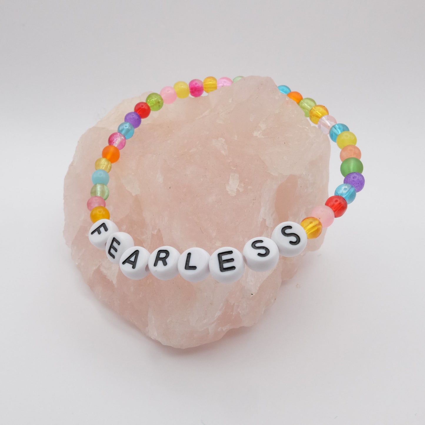 An elastic beaded bracelet composed of colourful 4mm glass beads in red, orange, yellow, green, aqua, blue, purple and pink with white acrylic letter beads spelling “FEARLESS,” displayed on a pale pink crystal rock.