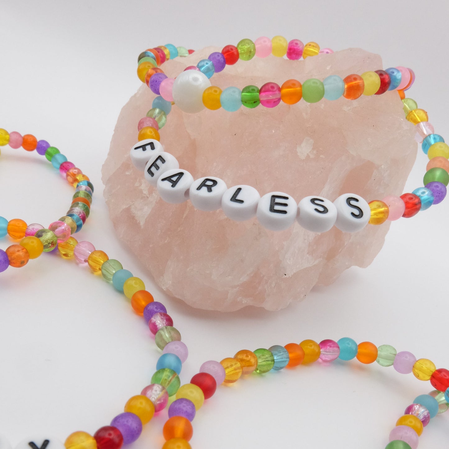 A group of elastic beaded bracelets composed of colourful 4mm glass beads in red, orange, yellow, green, aqua, blue, purple and pink with white acrylic letter beads spelling “FEARLESS,” displayed on a pale pink crystal rock.