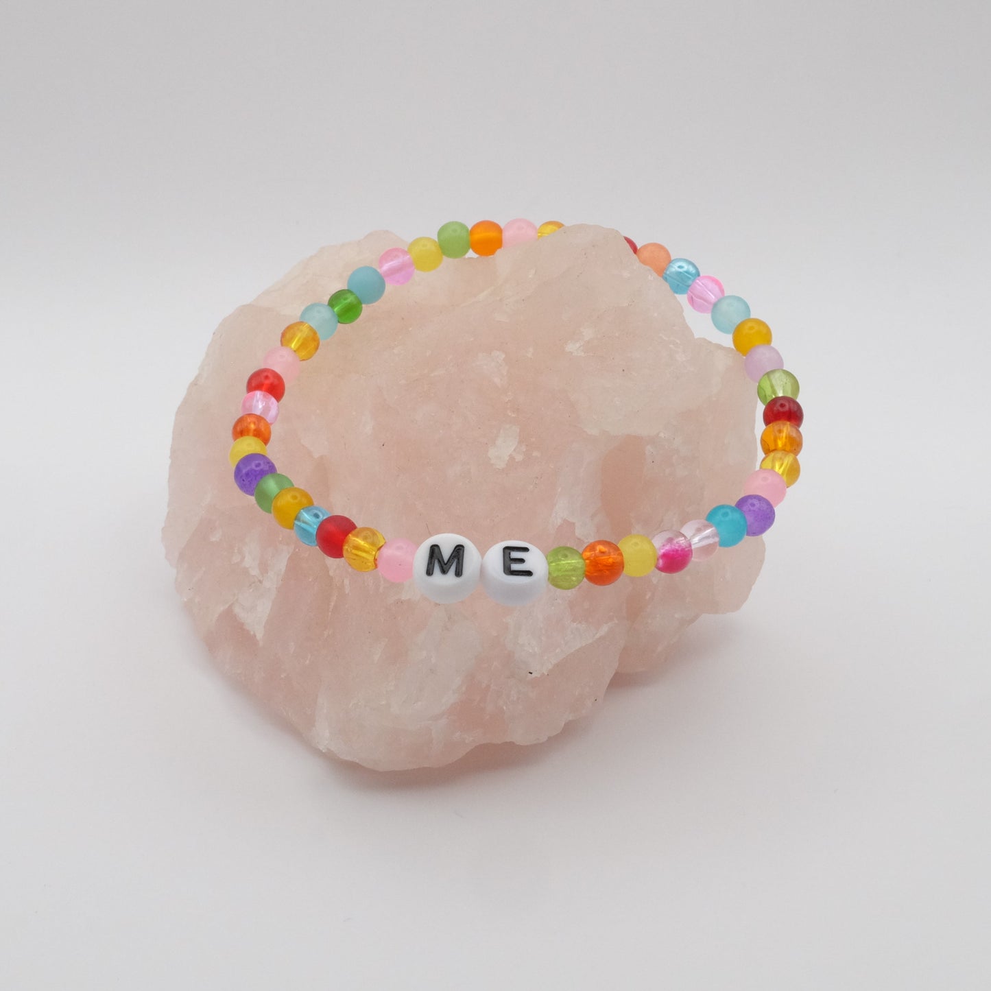 An elastic beaded bracelet composed of colourful 4mm glass beads in red, orange, yellow, green, aqua, blue, purple and pink with white acrylic letter beads spelling “ME,” displayed on a pale pink crystal rock.