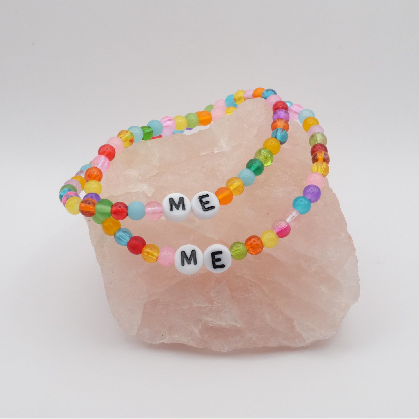 Two elastic beaded bracelets composed of colourful 4mm glass beads in red, orange, yellow, green, aqua, blue, purple and pink with white acrylic letter beads spelling “ME,” displayed on a pale pink crystal rock.