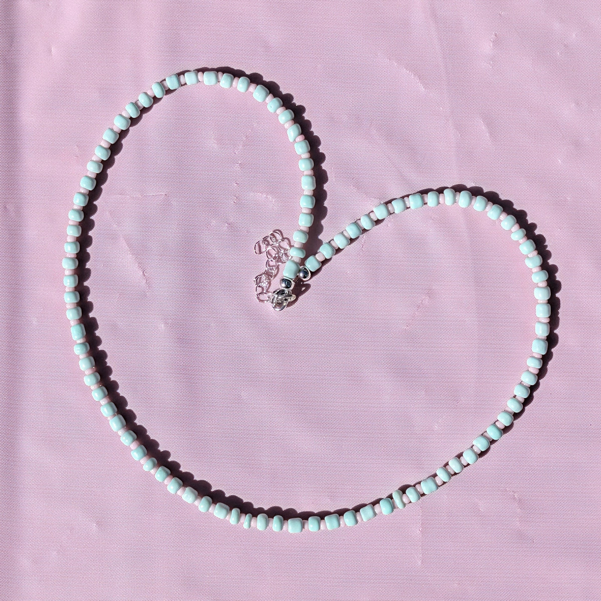 A heart-shaped beaded necklace made of mint green and white beads with a silver-toned heart charm, laid flat on a textured pink background.