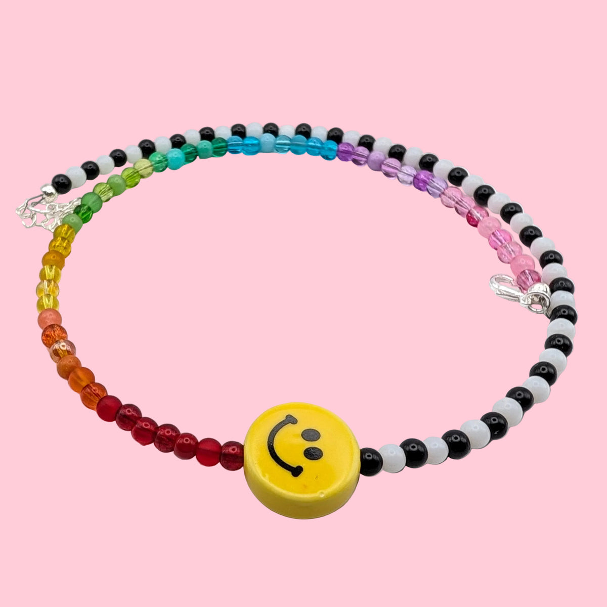 Tilly handmade necklace with rainbow and alternating black and white glass beads, centred with a yellow ceramic smiley face on a pink background.