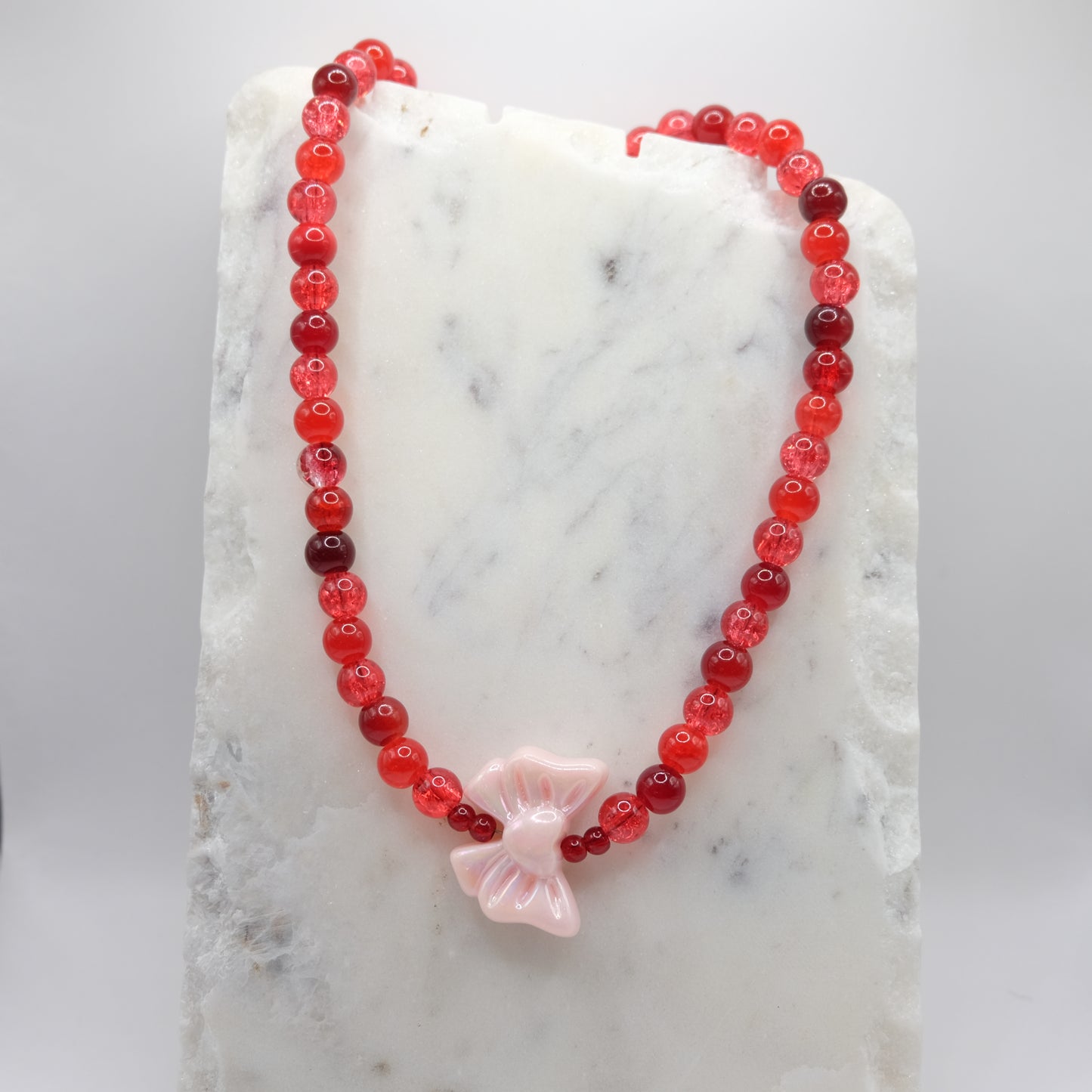 A beaded necklace composed of 8mm red glass beads - featuring an oversized pink acrylic bow bead with small red accents, shown on a white marble background.