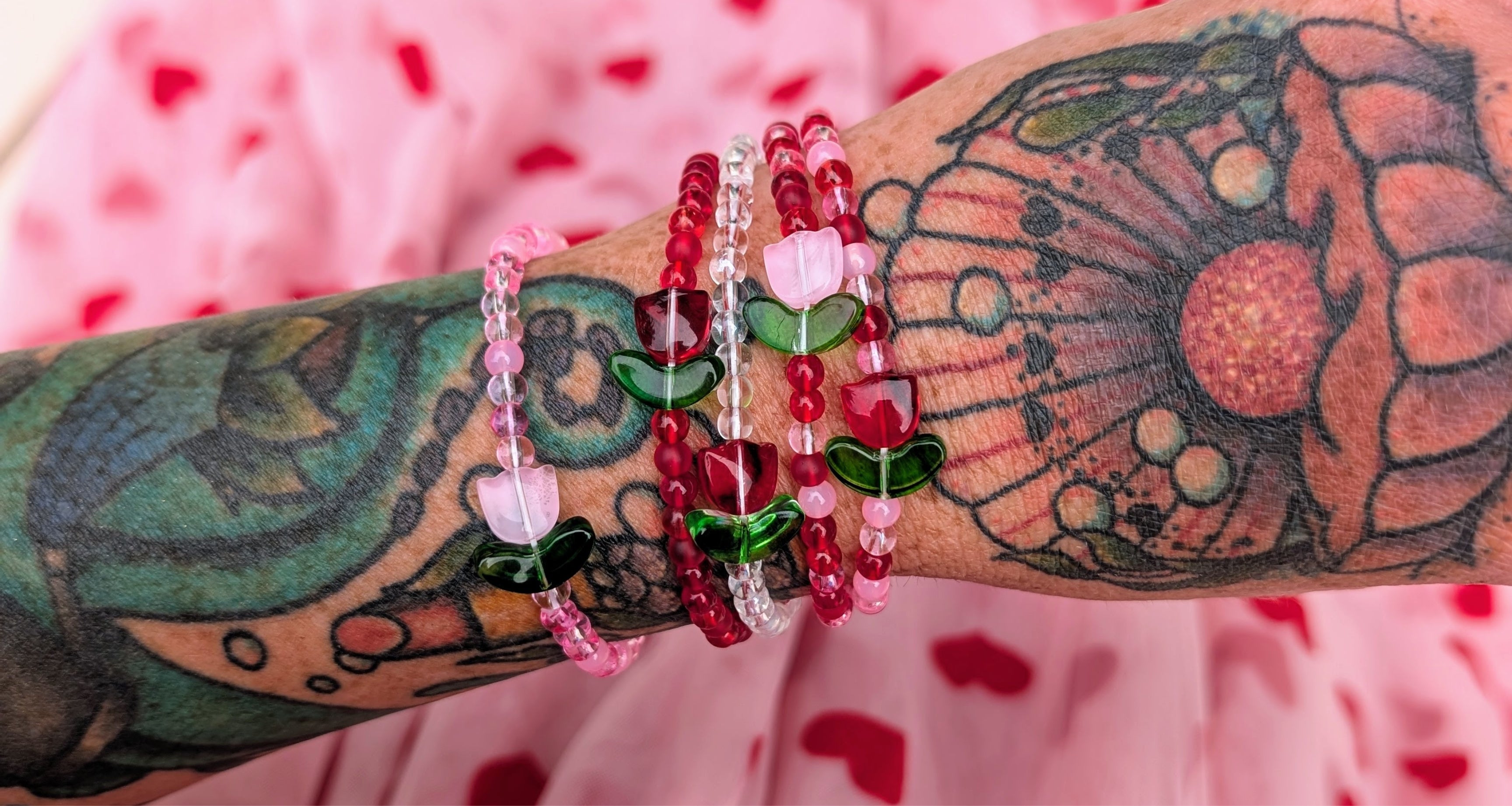 A tattooed arm wearing a stack of red and pink elastic beaded bracelets featuring tulip-shaped glass beads. Showcases handmade jewellery, perfect for dopamine dressing and quirky jewellery lovers. Displayed over a pink skirt with heart details.