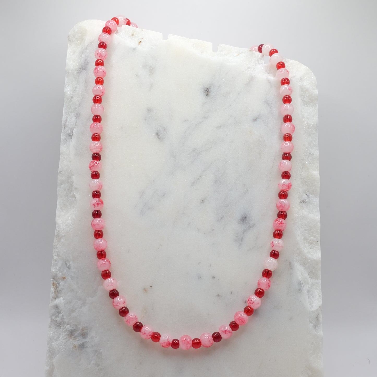 A beaded necklace composed of alternating 4mm and 6mm cherry red and marbled white and red glass beads - featuring glossy finishes, shown on a white marble background.