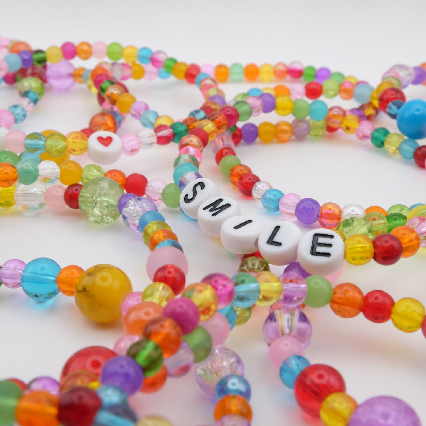 A flat lay of elastic beaded bracelets composed of vibrant 4mm glass beads in red, orange, yellow, green, aqua, blue, purple and pink with white acrylic letter beads spelling “SMILE,” displayed on a pale pink crystal rock.