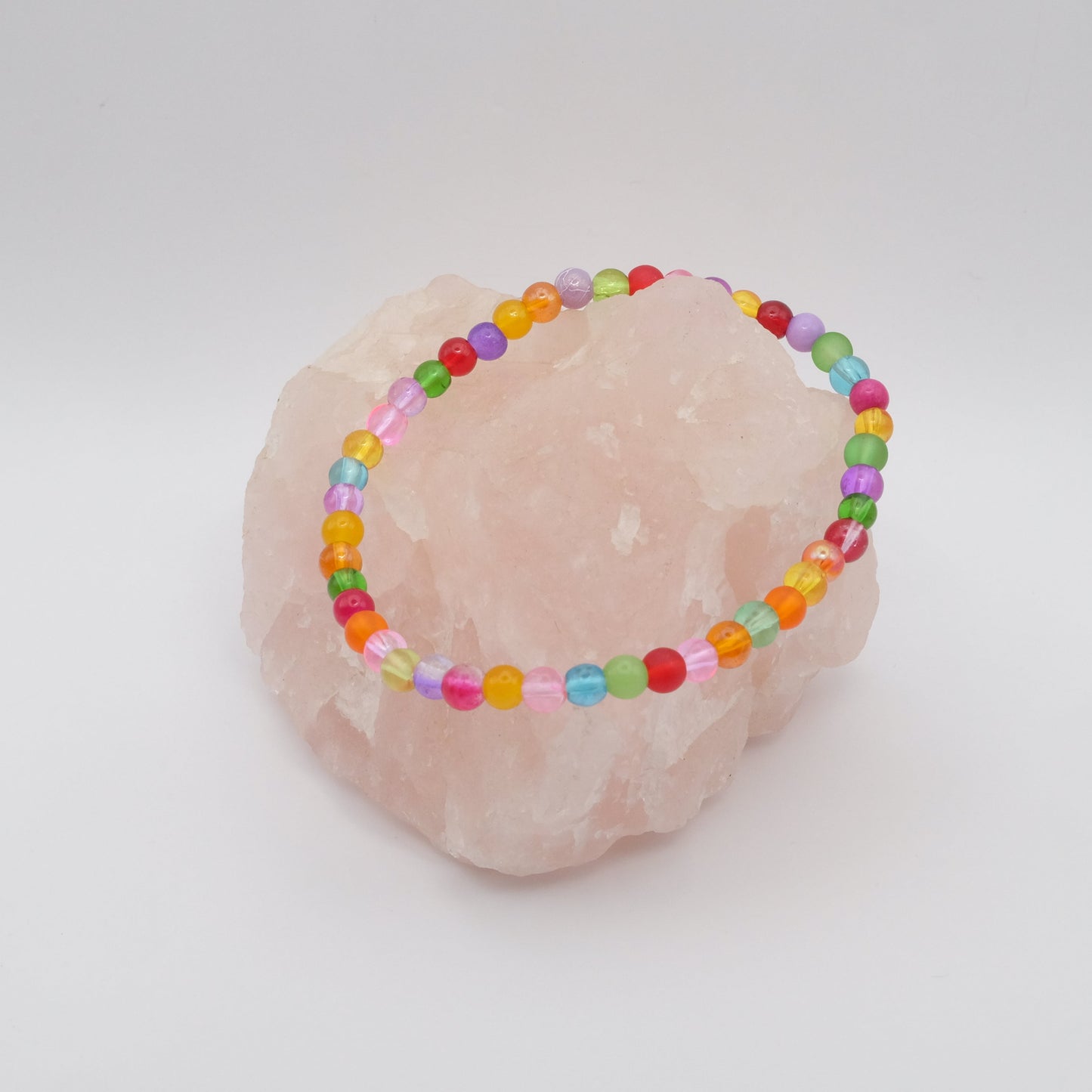 An elastic beaded bracelet composed of colourful 4mm glass beads in red, orange, yellow, green, aqua, blue, purple and pink, displayed on a pale pink crystal rock.