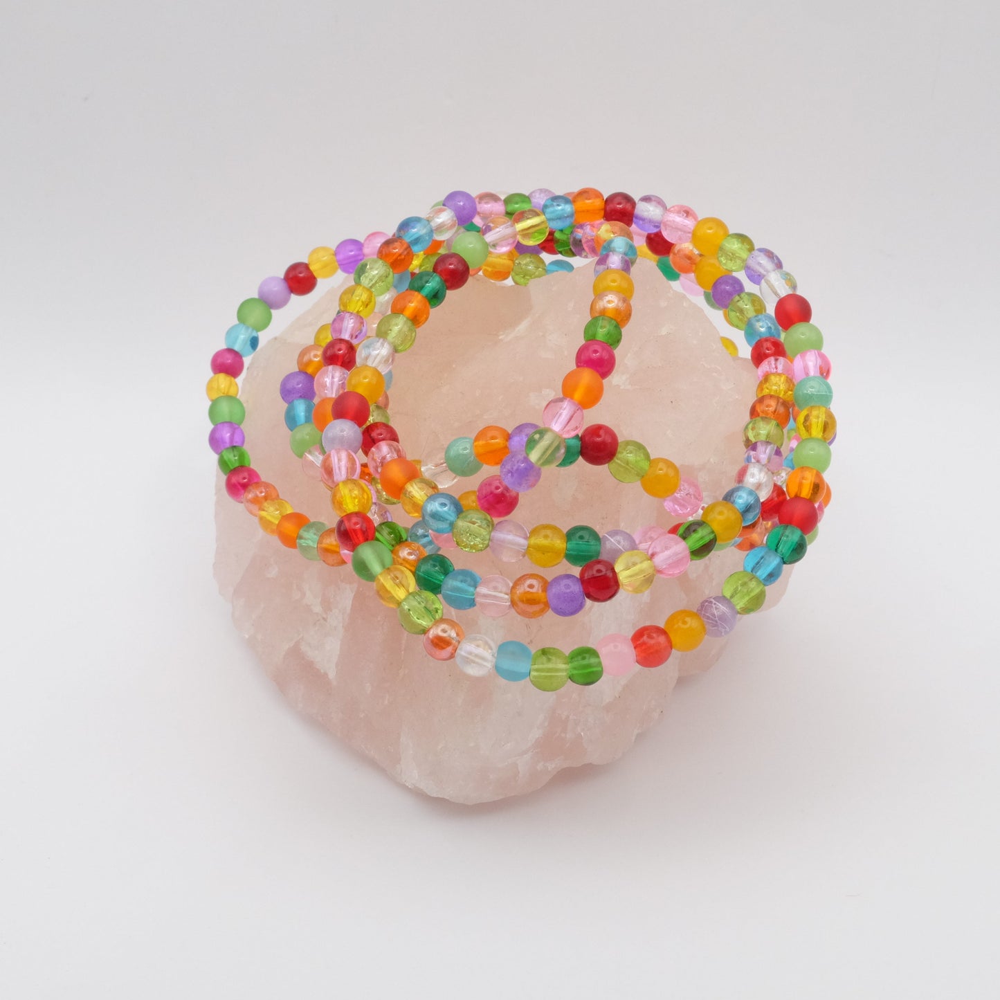 A group of elastic beaded bracelets composed of colourful 4mm glass beads in red, orange, yellow, green, aqua, blue, purple and pink, displayed on a pale pink crystal rock.