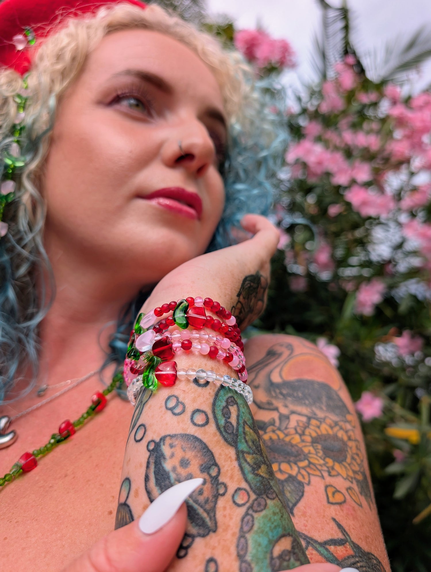 A woman with blue curls and floral tattoos wears a stack of red and pink elastic beaded bracelets made with glass beads, highlighting handmade jewellery and dopamine dressing. Set against a backdrop of pink flowers.
