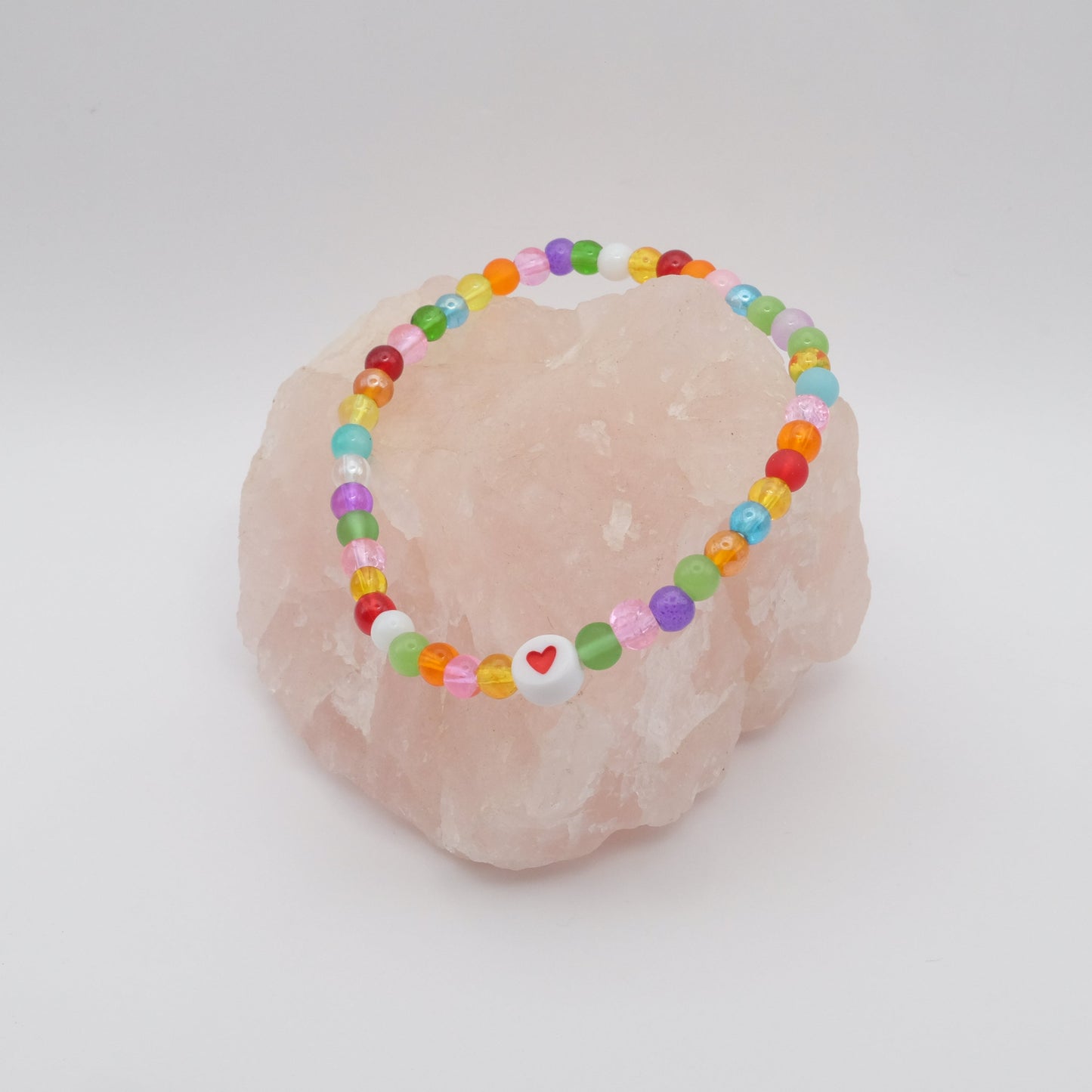 An elastic beaded bracelet composed of colourful 4mm glass beads in red, orange, yellow, green, aqua, blue, purple and pink with white and red acrylic love heart bead, displayed on a pale pink ring holder.