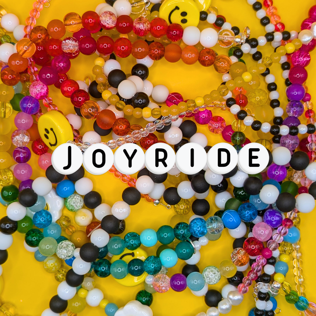 A flat lay of colourful handmade beaded jewellery on a bright yellow background featuring rainbow glass beads, black and white beads, smiley faces, freshwater pearls and layered textures, with “JOYRIDE” spelled in white letter beads at the centre.