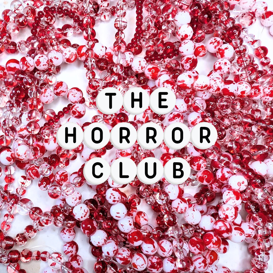 An assortment of elastic beaded bracelets composed of 8mm and 6mm glass beads in glossy white with vivid red splatter and clear glass with red splatter—arranged in a flat lay with white acrylic letter beads spelling “THE HORROR CLUB.”