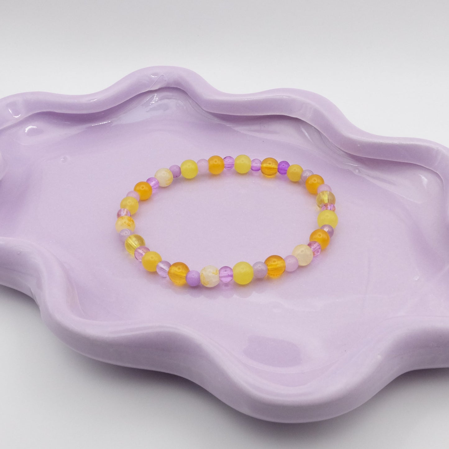 An elastic beaded bracelet featuring alternating 6mm yellow glass beads and 4mm purple glass beads in various shades, resting on a lavender wavy-edged dish against a light background.