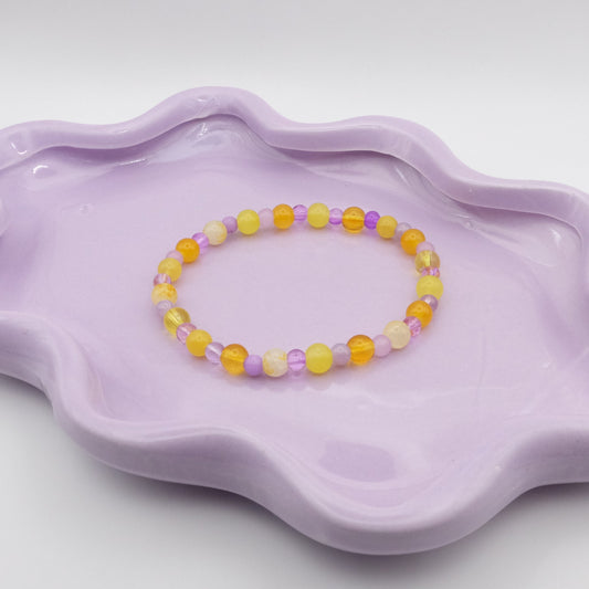 An elastic beaded bracelet featuring alternating 6mm yellow glass beads and 4mm purple glass beads in various shades, resting on a lavender wavy-edged dish against a light background.