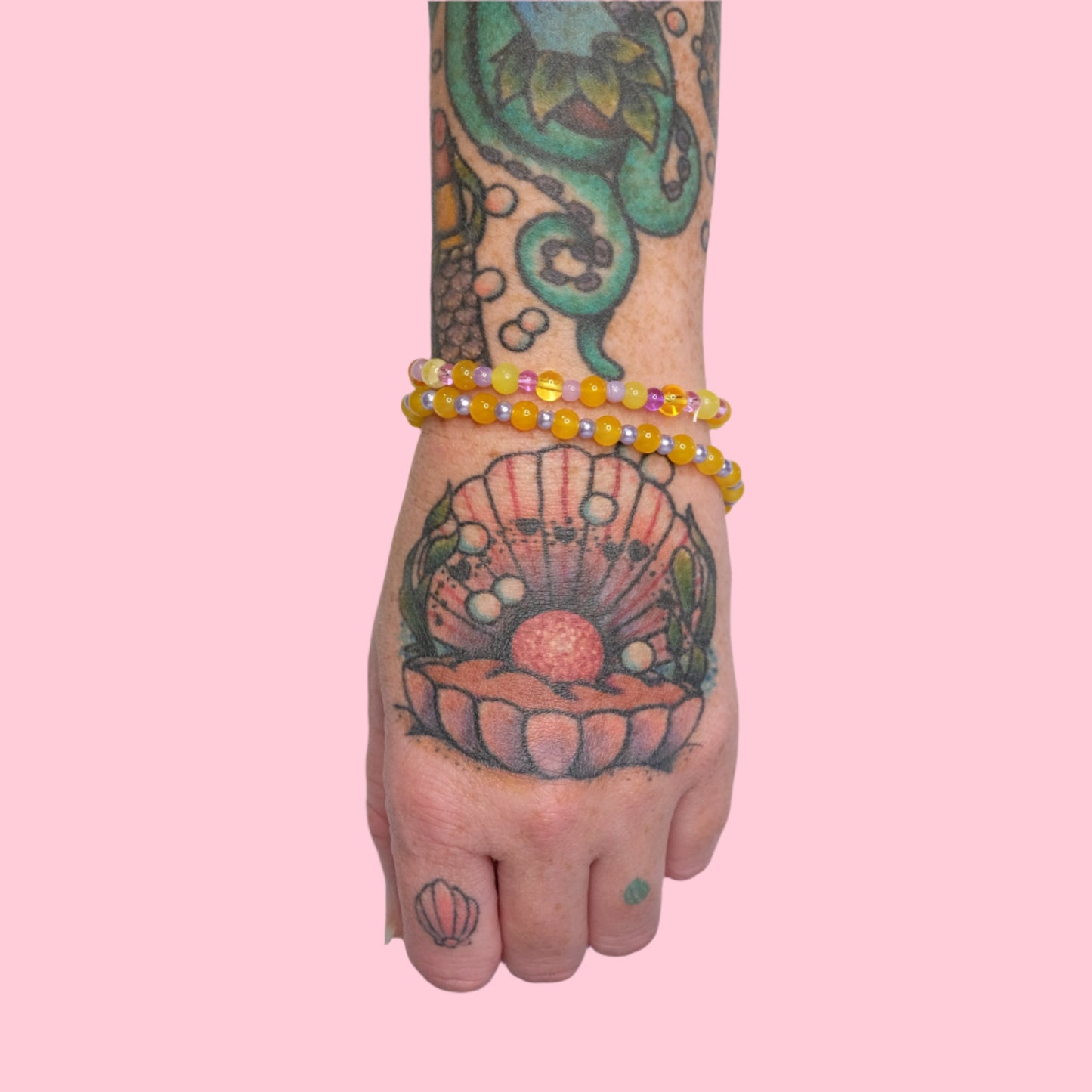 An arm decorated with sea-life tattoos, sporting multiple elastic beaded bracelets in soft purples and sunny yellows, set against a vibrant pink background.