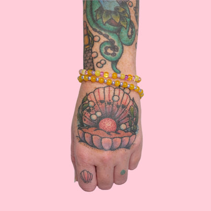 An arm decorated with sea-life tattoos, sporting multiple elastic beaded bracelets in soft purples and sunny yellows, set against a vibrant pink background.