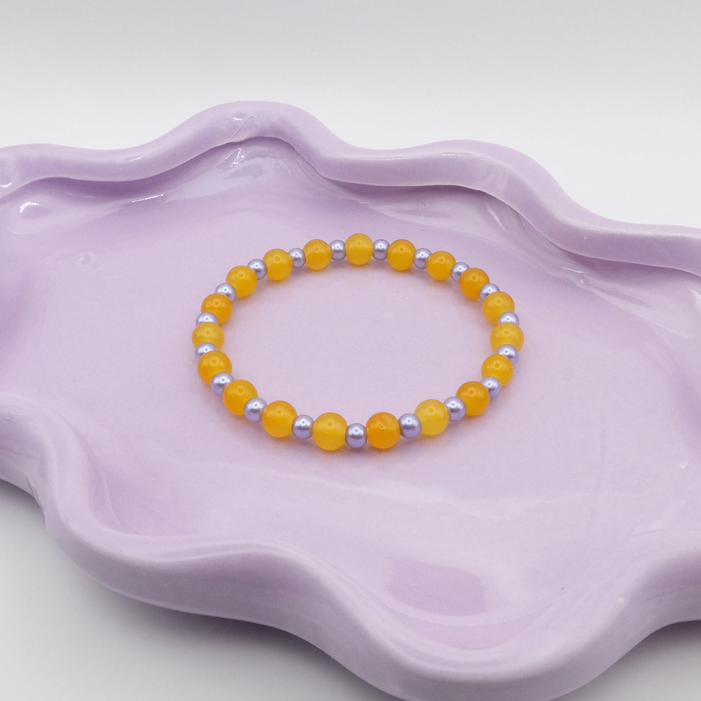 An elastic beaded bracelet showcasing alternating 6mm bright yellow glass beads paired with 4mm metallic purple beads, displayed on a lavender wavy-edged dish against a clean, neutral setting.