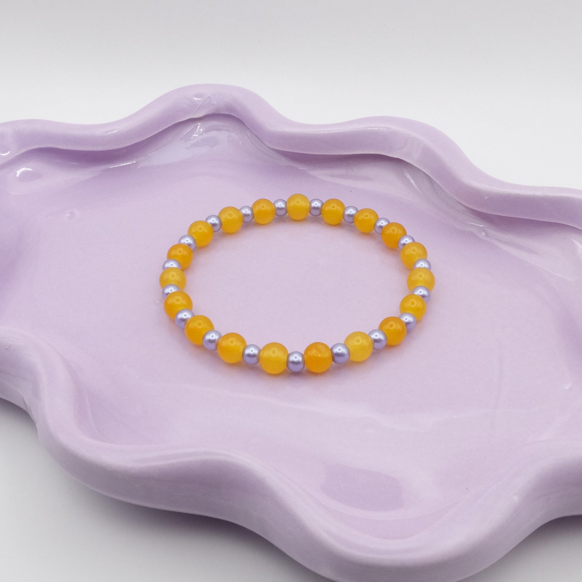 An elastic beaded bracelet showcasing alternating 6mm bright yellow glass beads paired with 4mm metallic purple beads, displayed on a lavender wavy-edged dish against a clean, neutral setting.