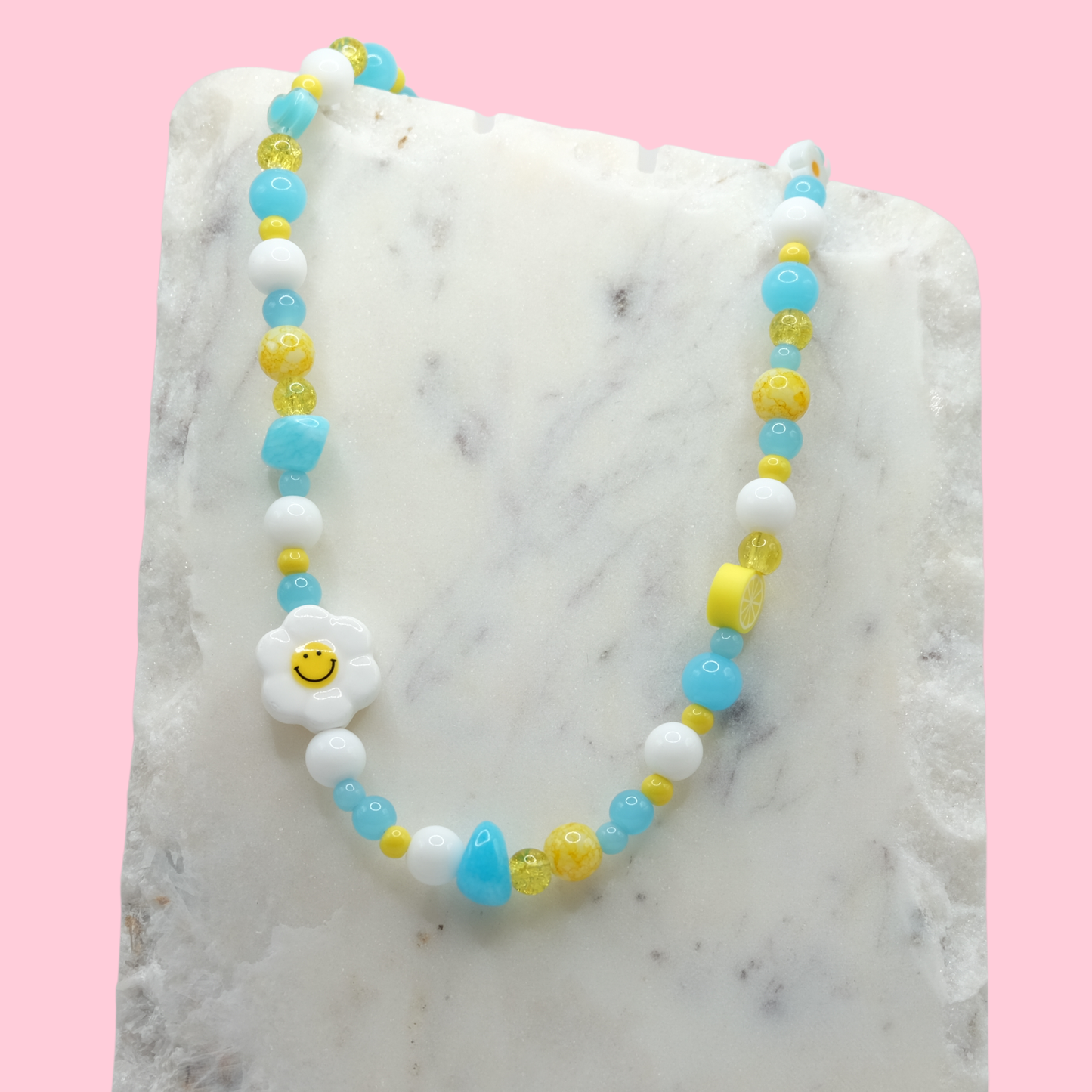 A handmade beaded necklace composed of mixed 4mm to 8mm blue, white, and yellow beads with blue dyed jade accents, featuring a ceramic daisy smiley face bead and a polymer clay lemon bead, displayed on a white marble slab.