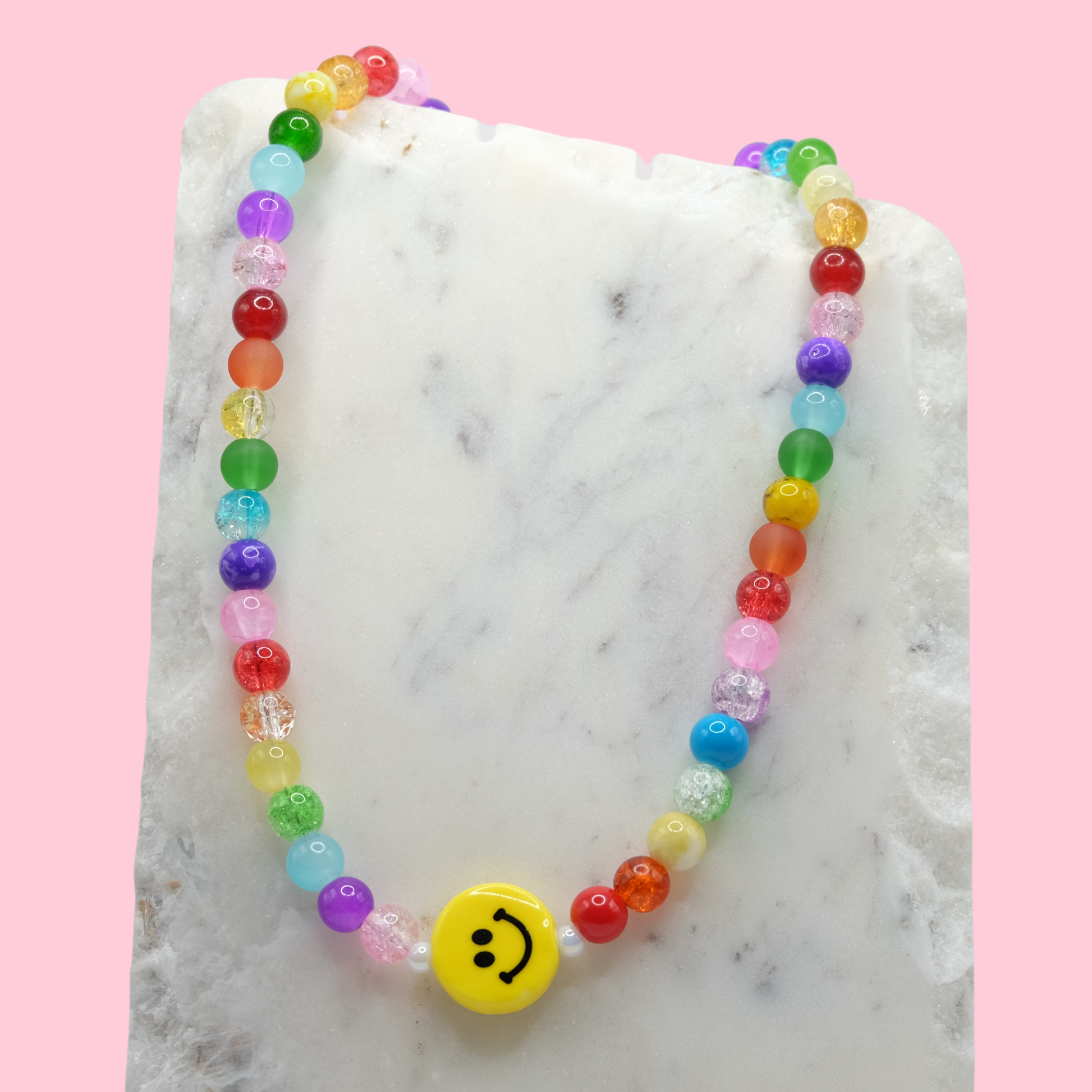 A handmade beaded necklace composed of rainbow-toned 8mm glass beads in glossy, frosted, and crackle finishes, featuring a yellow ceramic smiley face bead at the centre, displayed on a white marble slab.