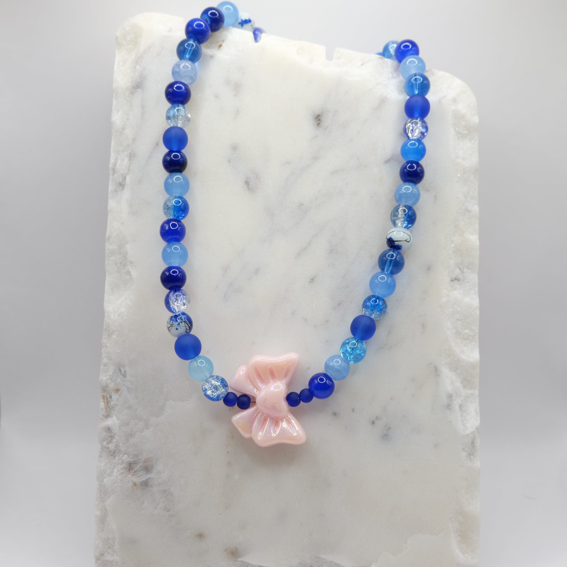 A beaded necklace composed of 8mm blue glass beads - featuring a large pink acrylic bow bead at the centre, shown on a white marble background.