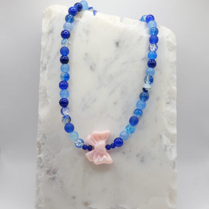 A beaded necklace composed of 8mm blue glass beads - featuring a large pink acrylic bow bead at the centre, shown on a white marble background.