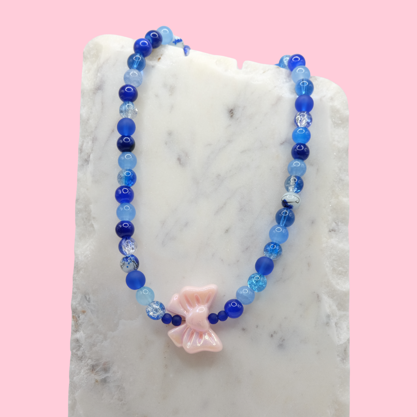 Blue beaded necklace with an oversized pink acrylic bow bead on a marble surface with a pink background