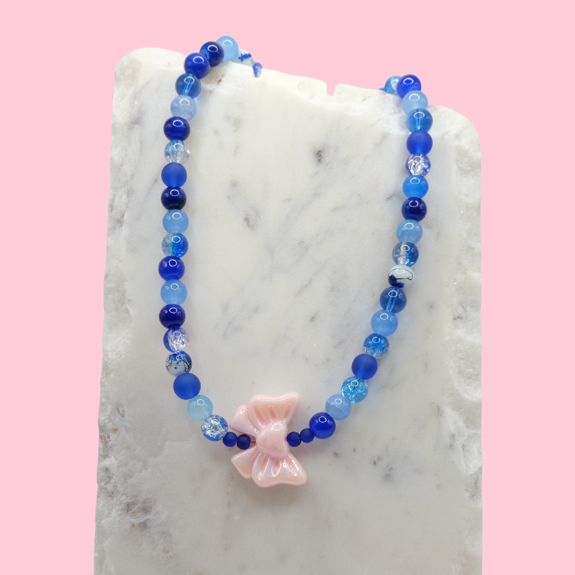 Blue beaded necklace with an oversized pink acrylic bow bead on a marble surface with a pink background