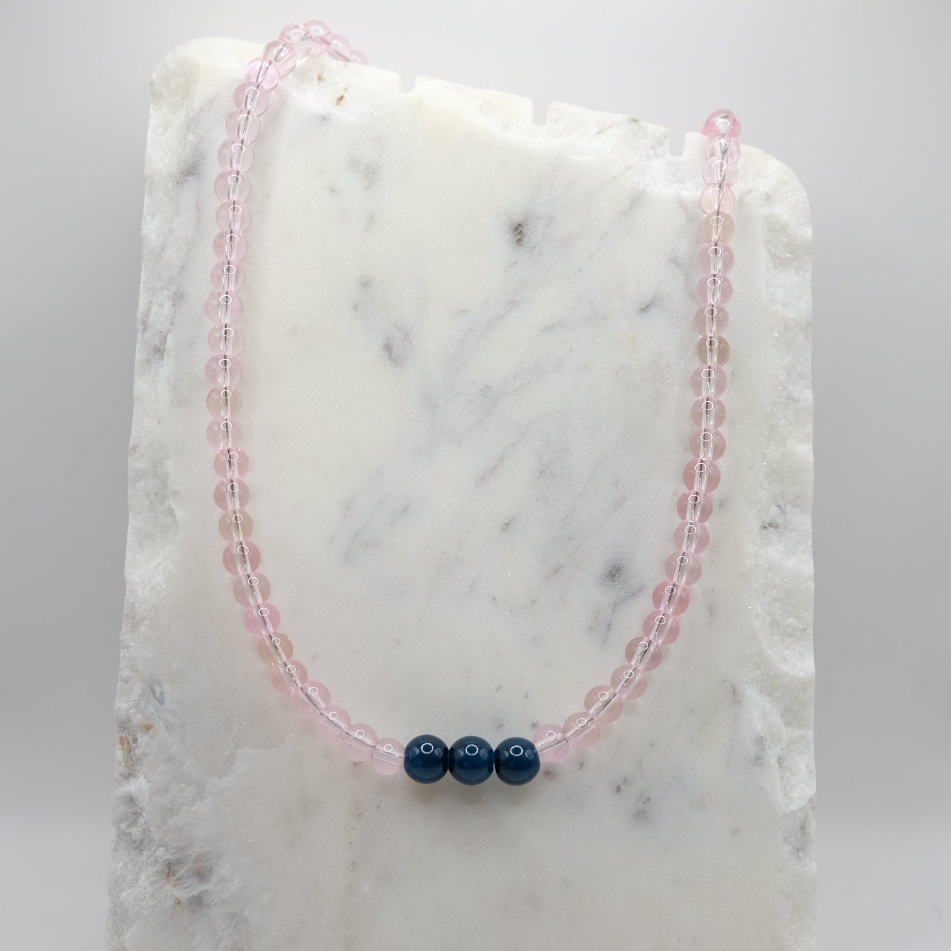 A beaded necklace composed of 6mm pale pink clear glass beads - featuring three 8mm navy blue glass beads at the centre, shown on a white marble background.