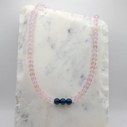 A beaded necklace composed of 6mm pale pink clear glass beads - featuring three 8mm navy blue glass beads at the centre, shown on a white marble background.
