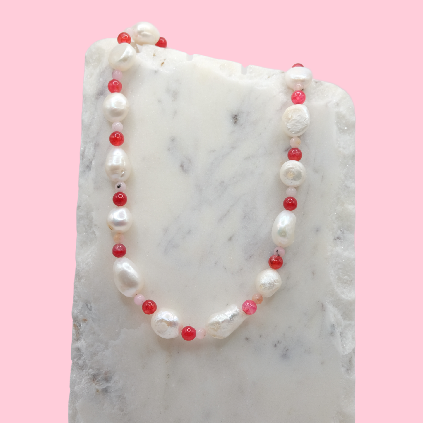 Necklace with alternating freshwater pearls and red glass beads on a marble surface with a pink background