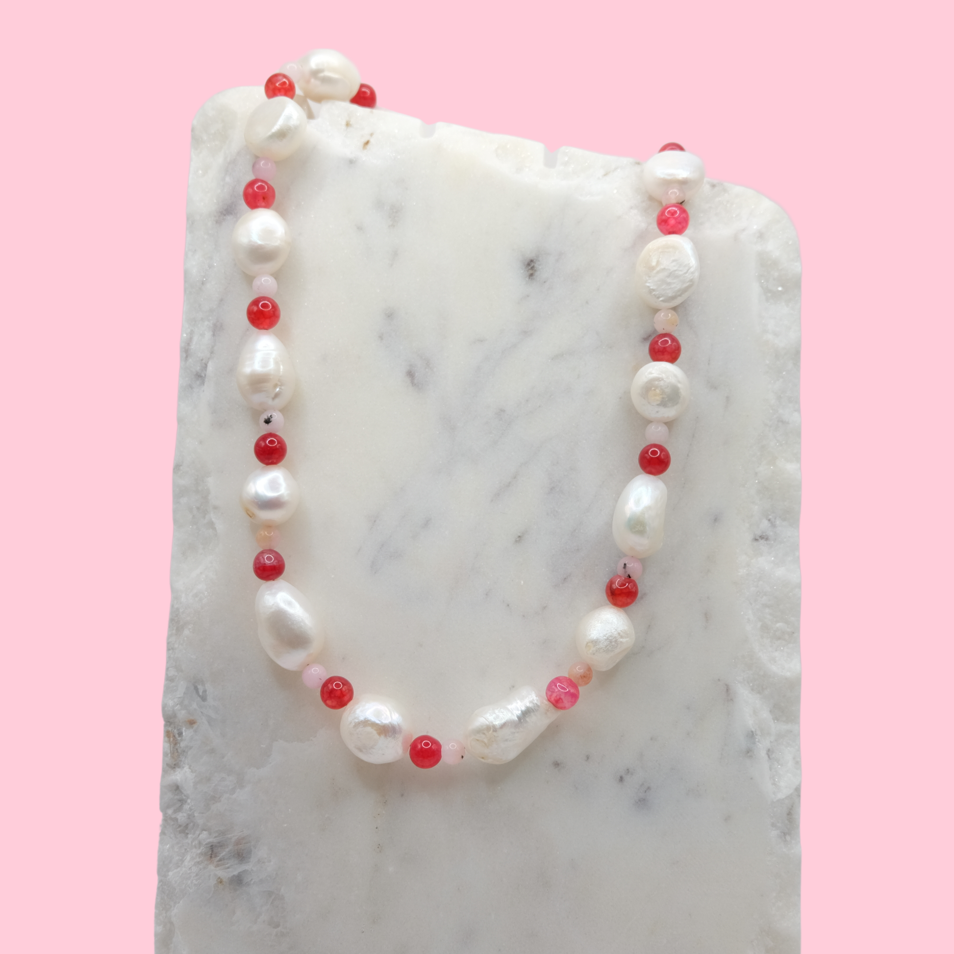 Necklace with alternating freshwater pearls and red glass beads on a marble surface with a pink background