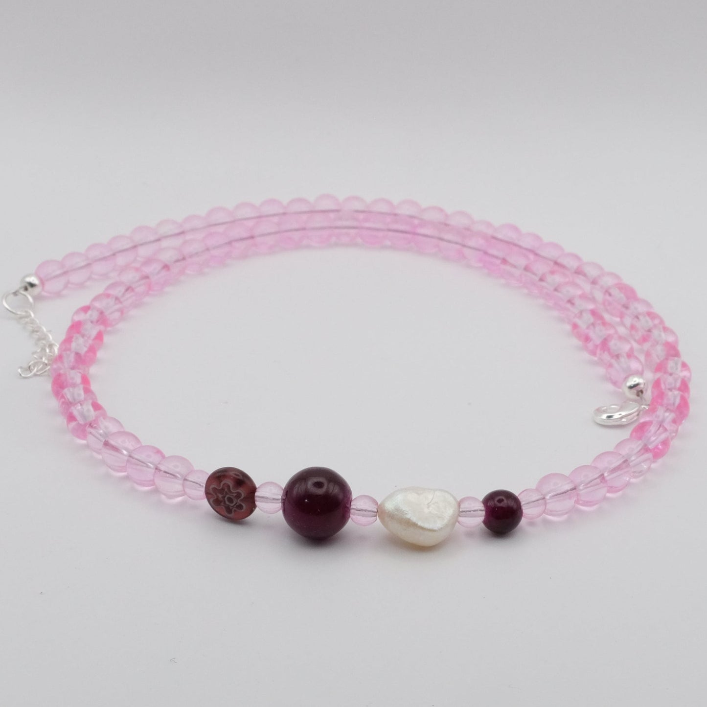 A handmade beaded necklace featuring translucent pink 6mm glass beads, plum glass feature beads and a freshwater pearl, displayed on a white marble stand.