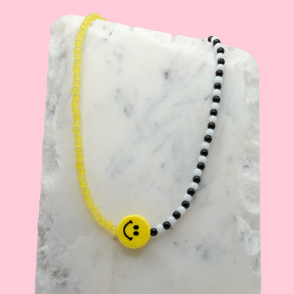 A handmade beaded necklace composed of yellow, black, and white 4mm glass beads, featuring a yellow ceramic smiley face bead at the centre, displayed on a white marble slab.