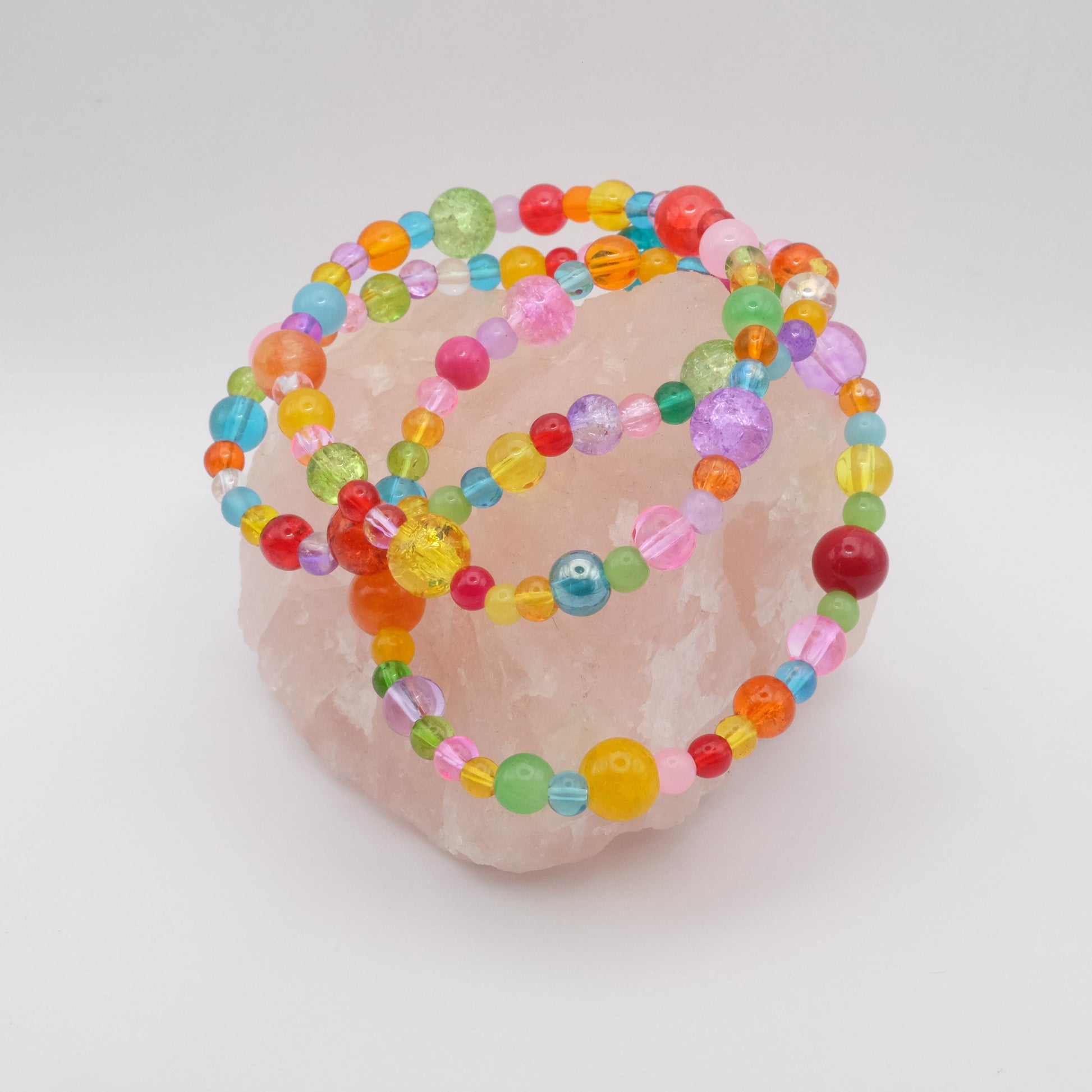 Three elastic beaded bracelets composed of assorted colourful 4mm,6mm and 8mm  glass beads in red, orange, yellow, green, aqua, blue, purple and pink - featuring frosted, transparent, and glossy textures, displayed on a pale pink crystal rock.