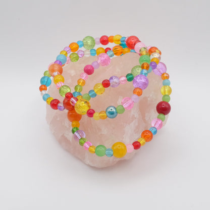 Three elastic beaded bracelets composed of assorted colourful 4mm,6mm and 8mm  glass beads in red, orange, yellow, green, aqua, blue, purple and pink - featuring frosted, transparent, and glossy textures, displayed on a pale pink crystal rock.