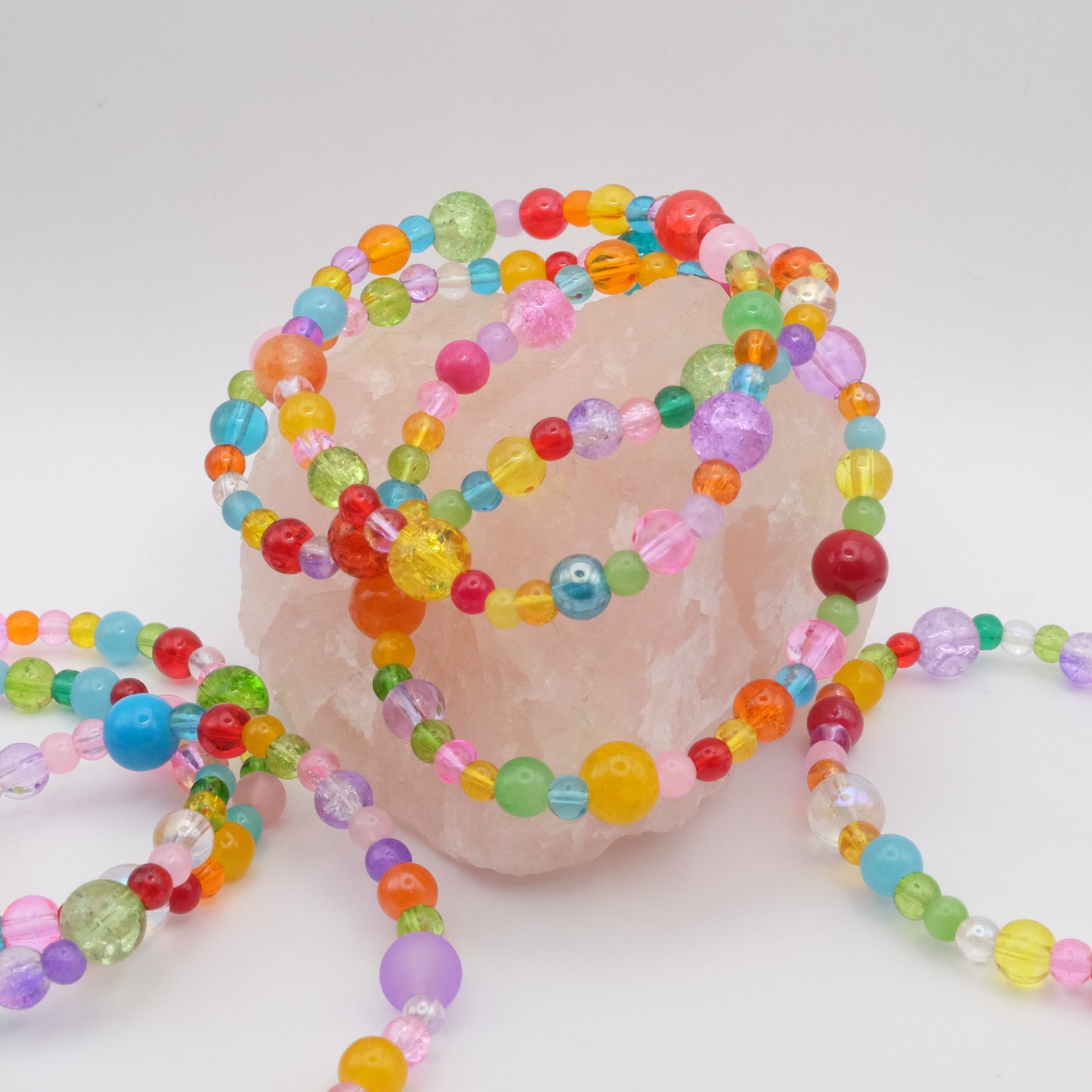Multiple elastic beaded bracelets composed of assorted colourful 4mm,6mm and 8mm  glass beads in red, orange, yellow, green, aqua, blue, purple and pink - featuring frosted, transparent, and glossy textures, displayed on a pale pink crystal rock.