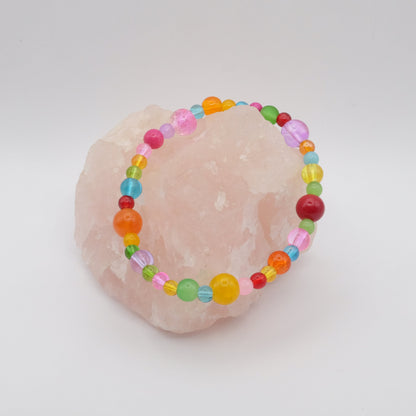 An elastic beaded bracelet composed of assorted colourful 4mm,6mm and 8mm  glass beads in red, orange, yellow, green, aqua, blue, purple and pink - featuring frosted, transparent, and glossy textures, displayed on a pale pink crystal rock.