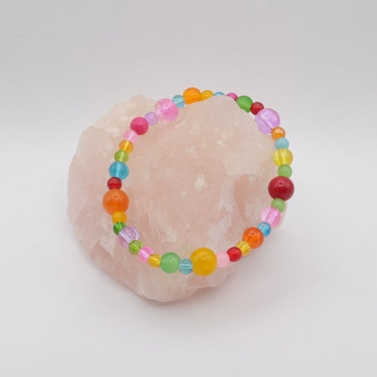 An elastic beaded bracelet composed of assorted colourful 4mm,6mm and 8mm  glass beads in red, orange, yellow, green, aqua, blue, purple and pink - featuring frosted, transparent, and glossy textures, displayed on a pale pink crystal rock.