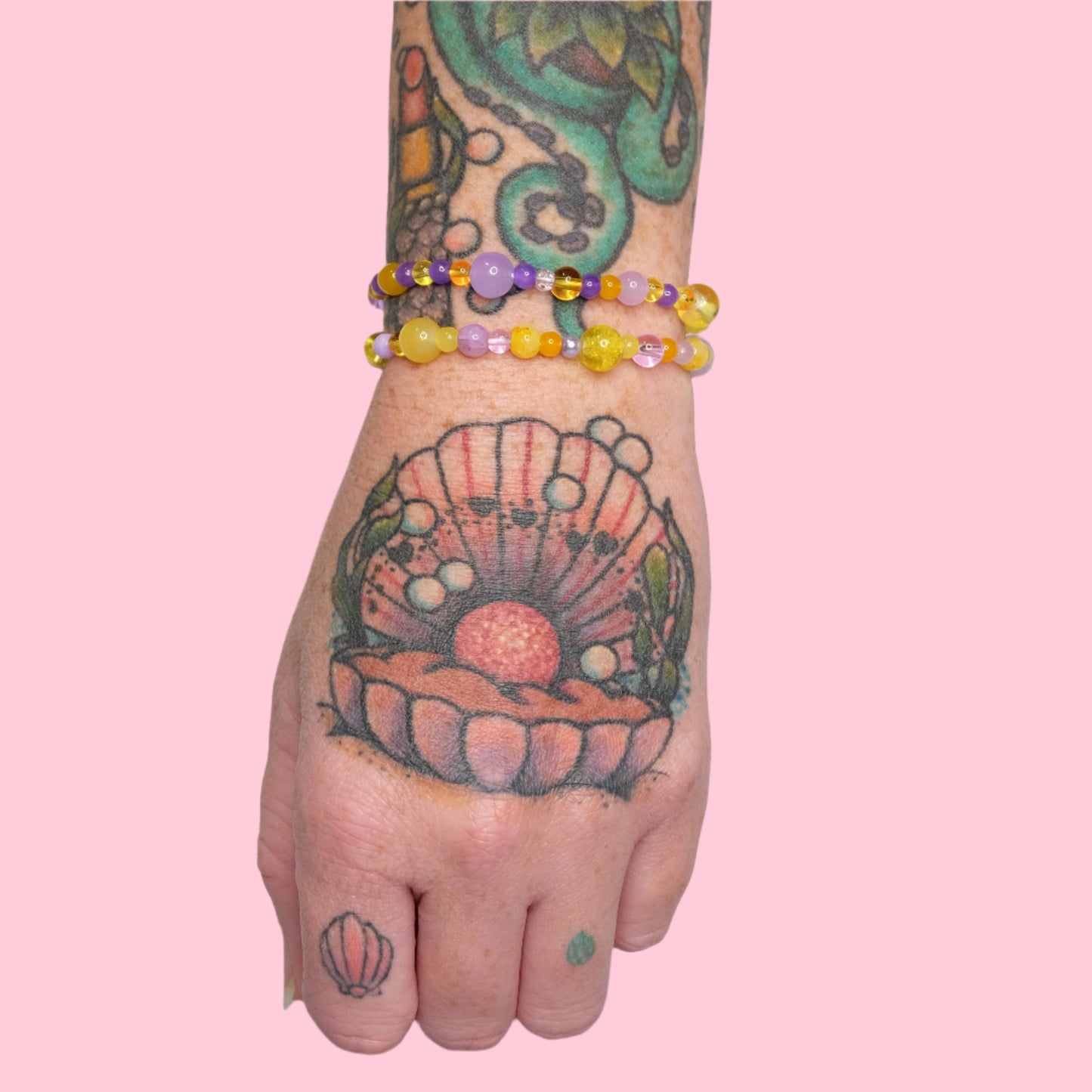 A tattooed arm with octopus and seashell tattoos, wearing two elastic beaded bracelets in yellow and lavender hues, set against a solid pink background.