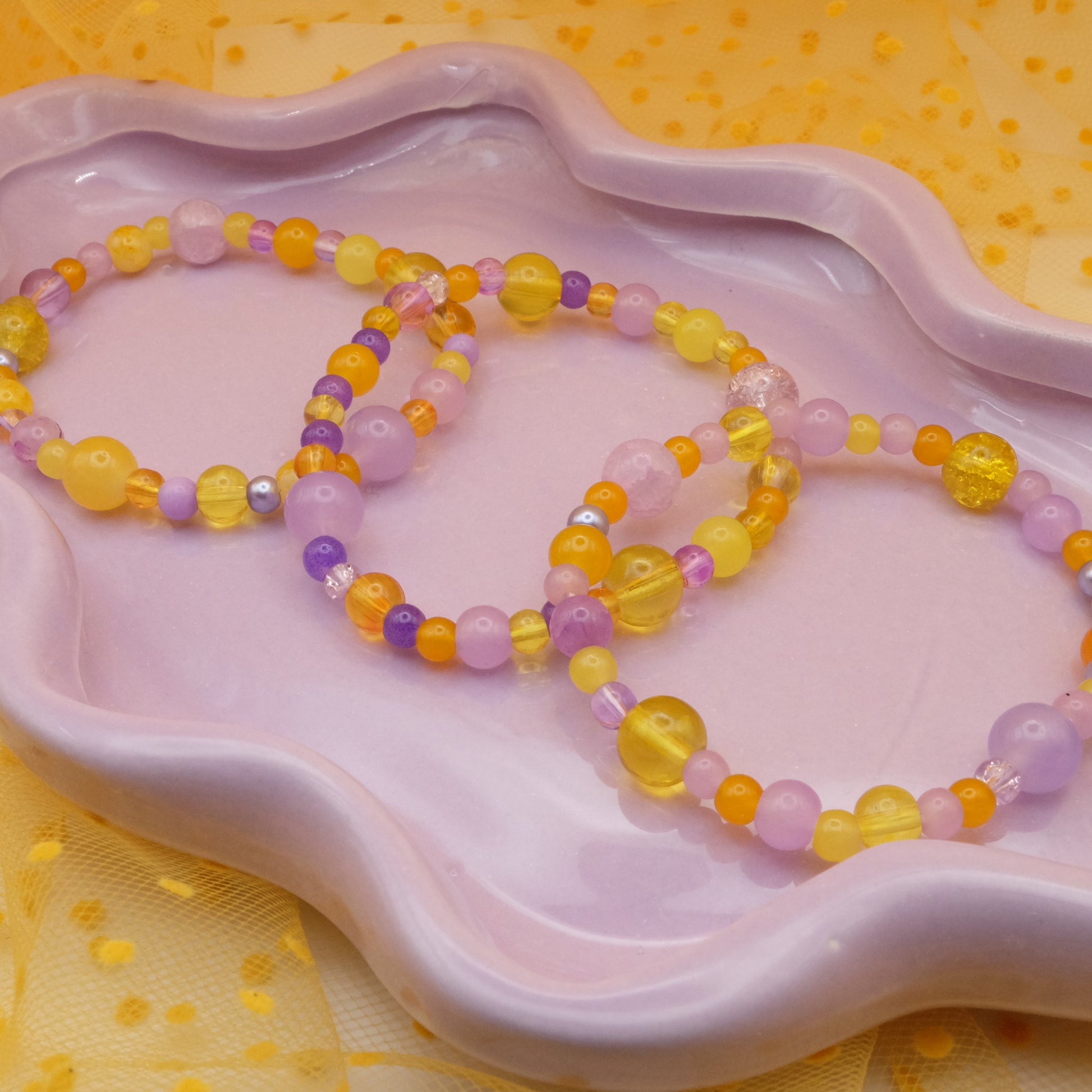 Three elastic beaded bracelets featuring 4mm, 6mm, and 8mm glass beads in shades of purple and yellow arranged on a lavender dish with a soft polka-dot netting backdrop.