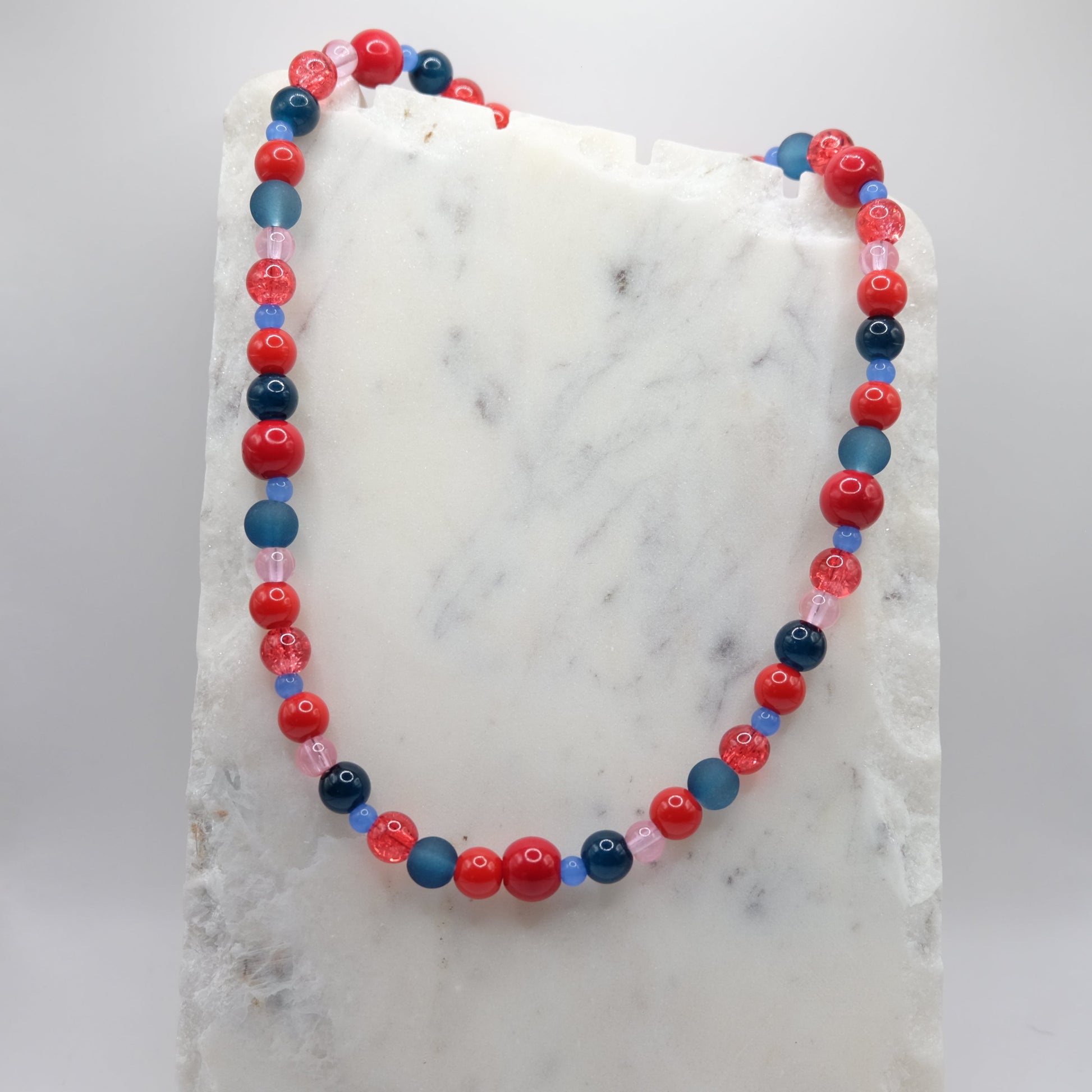 A beaded necklace composed of mixed 4mm, 6mm, 8mm and 10mm glass beads in red, blue and pink tones - featuring varied finishes, shown on a white marble background.
