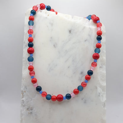 A beaded necklace composed of mixed 4mm, 6mm, 8mm and 10mm glass beads in red, blue and pink tones - featuring varied finishes, shown on a white marble background.